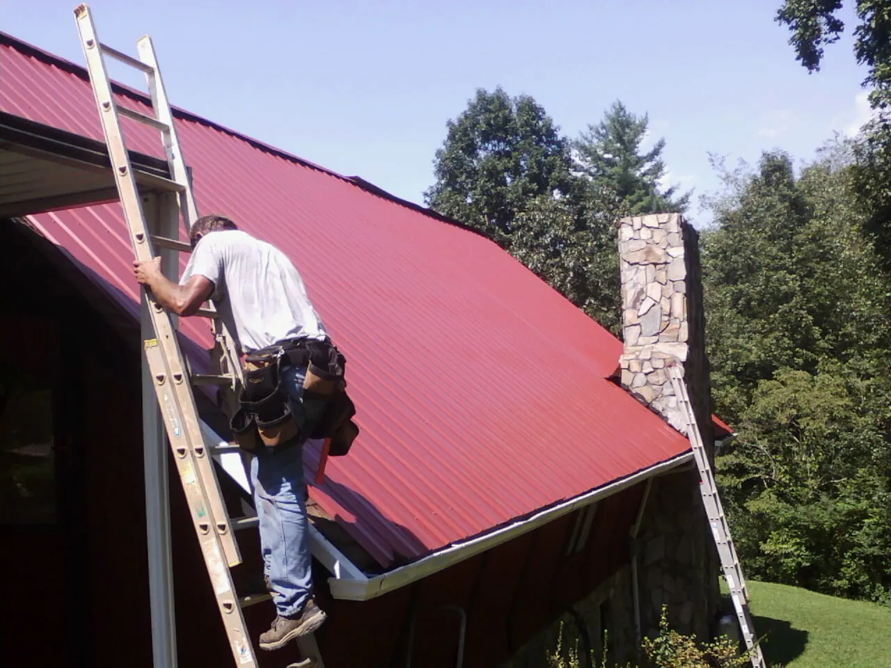 Roofer on a ladder beside a red metal roof and stone chimney — Soffit & Fascia Repair by Roofing Brothers in Cisco